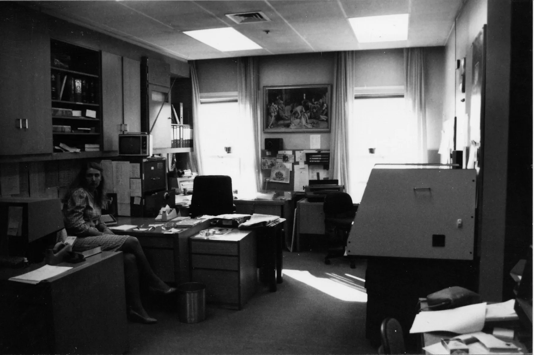 The black and white photograph shows Heather Mittag dressed in a plaid skirt suit sitting on a chair in a small open plan office. Several desks are dominated by large typewriters and computer monitors. The walls are filled with pinboards containing notes and reference sheets and there are numerous filing cabinets hold.  On the back wall is a framed print of the painting ‘The Shearing of the Rams’ by Tom Roberts.