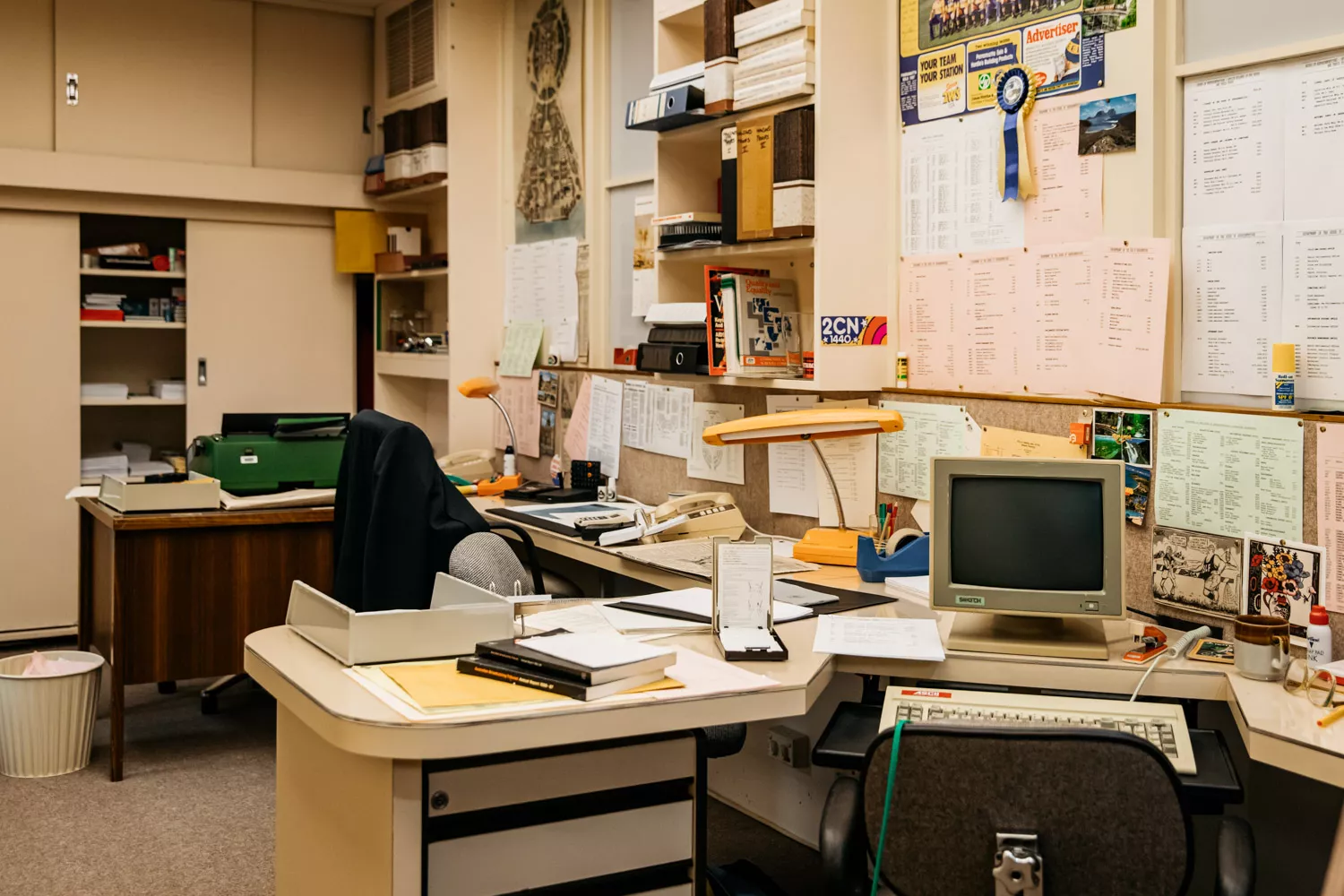 Two desks cluttered with books, pens, staplers, old fashioned 80s computers, and a pinboard covered in papers. 