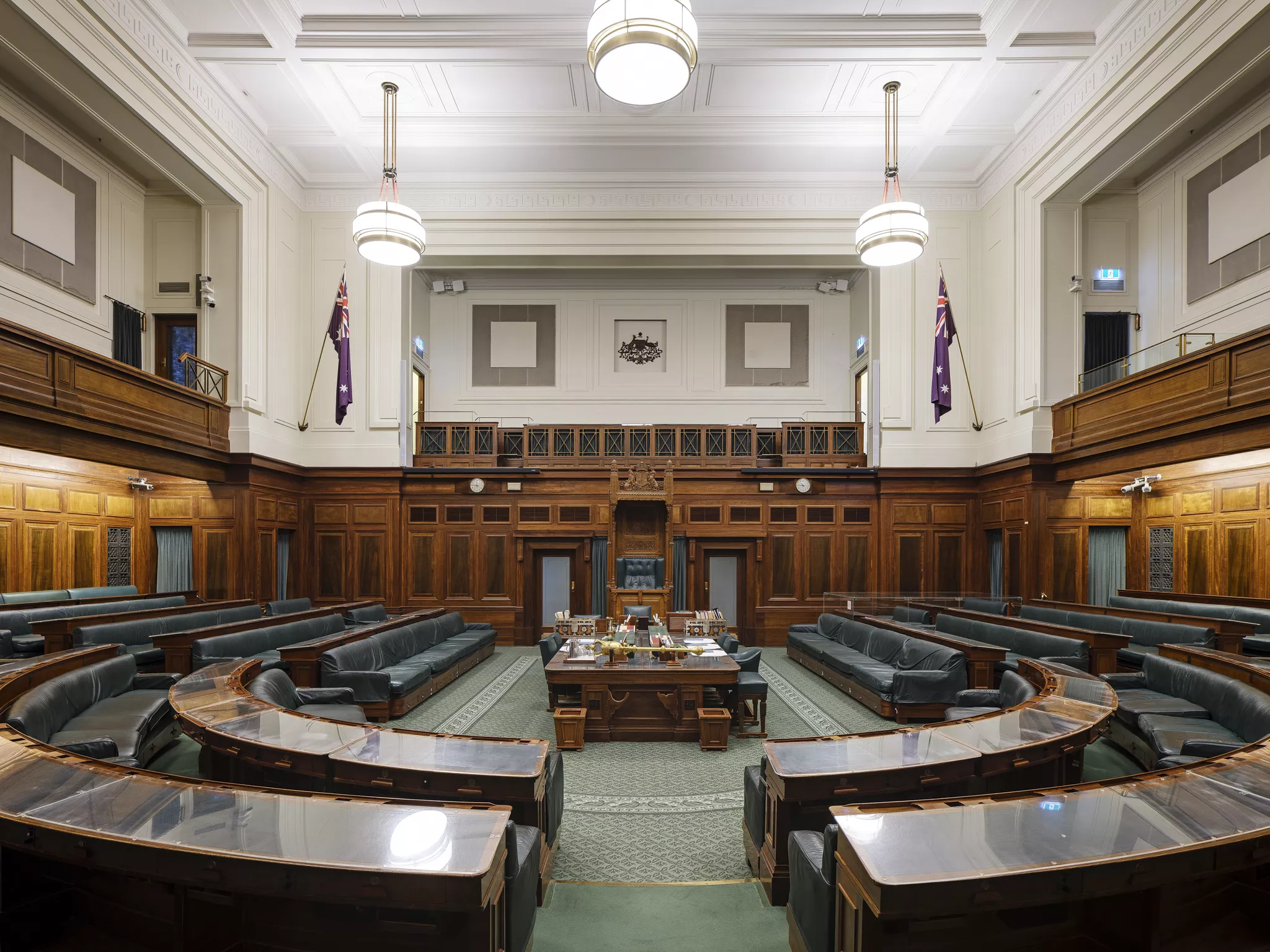 A green carpeted room with rows of wooden benches in a U shape facing a central table and the large, ornate Speaker's Chair. 