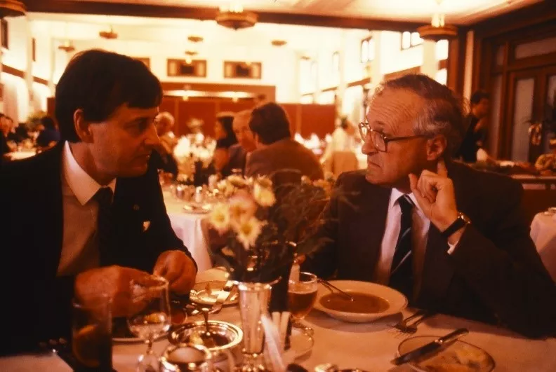 In this professional colour photograph, Gary Punch MP and Senator John Stone are seated at a dining table in the Members’ Dining Room midway through lunch. The table is laid with silverware, glassware and a floral arrangement. Gary Punch is wearing a dark suit with white shirt and striped tie. Senator Stone is similarly attired but in a brown suit and he is wearing glasses. In the background other politicians are dining and light is pouring in from high windows and French doors. 