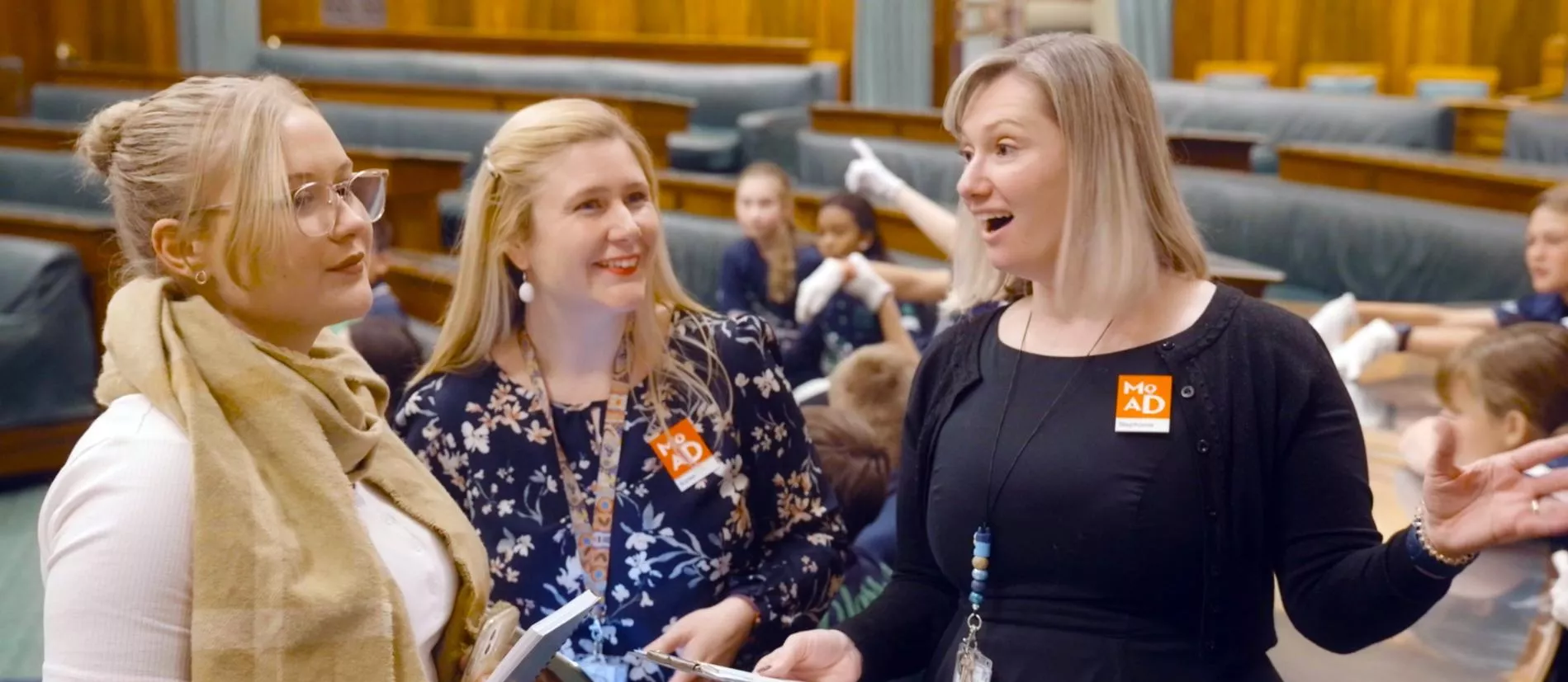 Two people stand holding clipboards, smiling and talking to a person about old parliament house's chamber of representatives.