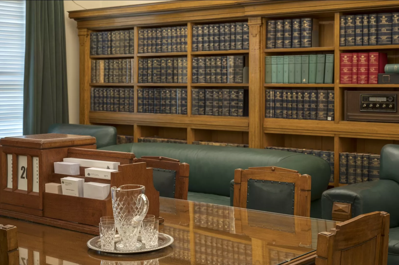 This colour photograph shows the Government Party Room re-created to evoke the atmosphere of 1964. There is a large meeting table in the foreground with a timber stationery holder and glass water jug. In the background are green leather easy chairs and settees and a large honey-coloured bookcase containing reference books, Hansards (volumes of parliamentary debates) and a radio. 