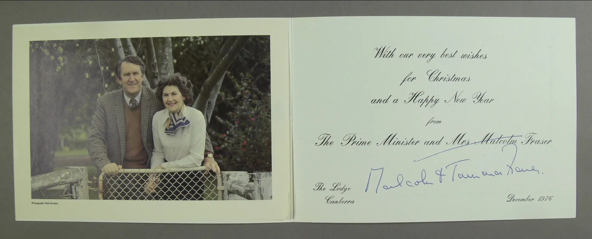 A christmas card featuring a photo of Malcolm Fraser wearing a tweed jacket and Tamie Fraser wearing a white blouse and blue striped scarf, posing together in from of a gate.