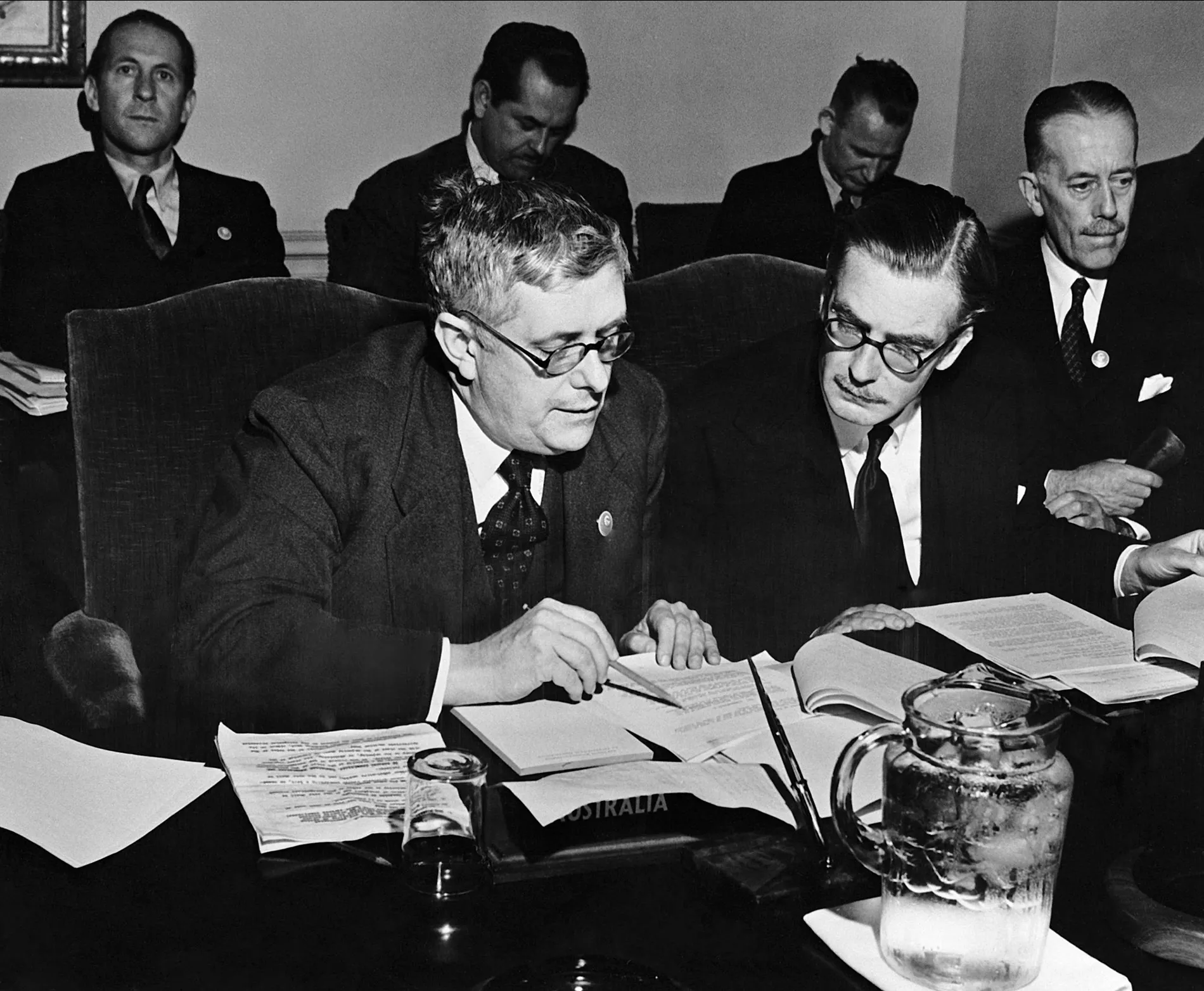 Dr Herbert Evatt sits next to Anthony Eden, reading through typed paper documents, at a table with a glass jug of iced water, and a nameplate labelled 'Australia'.