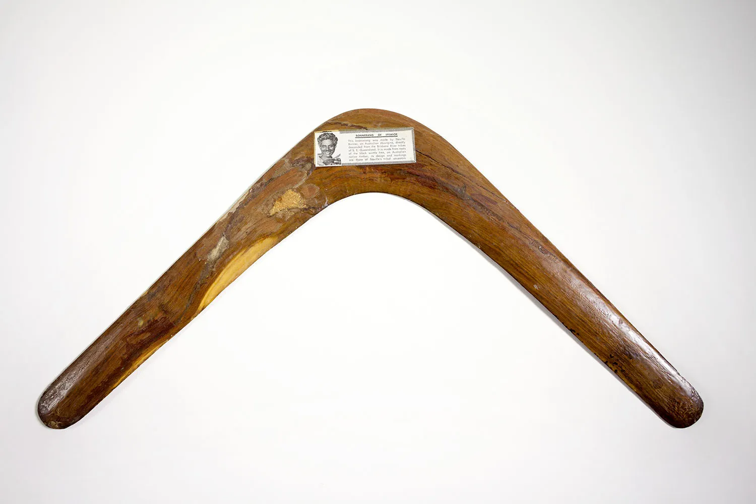 A wooden boomerang with a white descriptive label featuring text and a man's face.