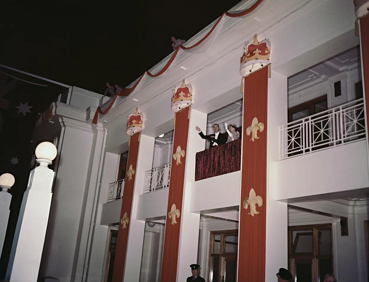 Prince Phillip and the Queen wave to a crowd from a balcony at Parliament House. It is at night.