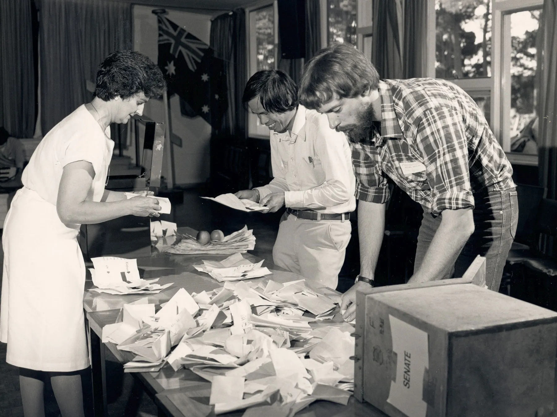 Three people stand around a table counting paper votes. There is a box titled 'Senate' on the table.