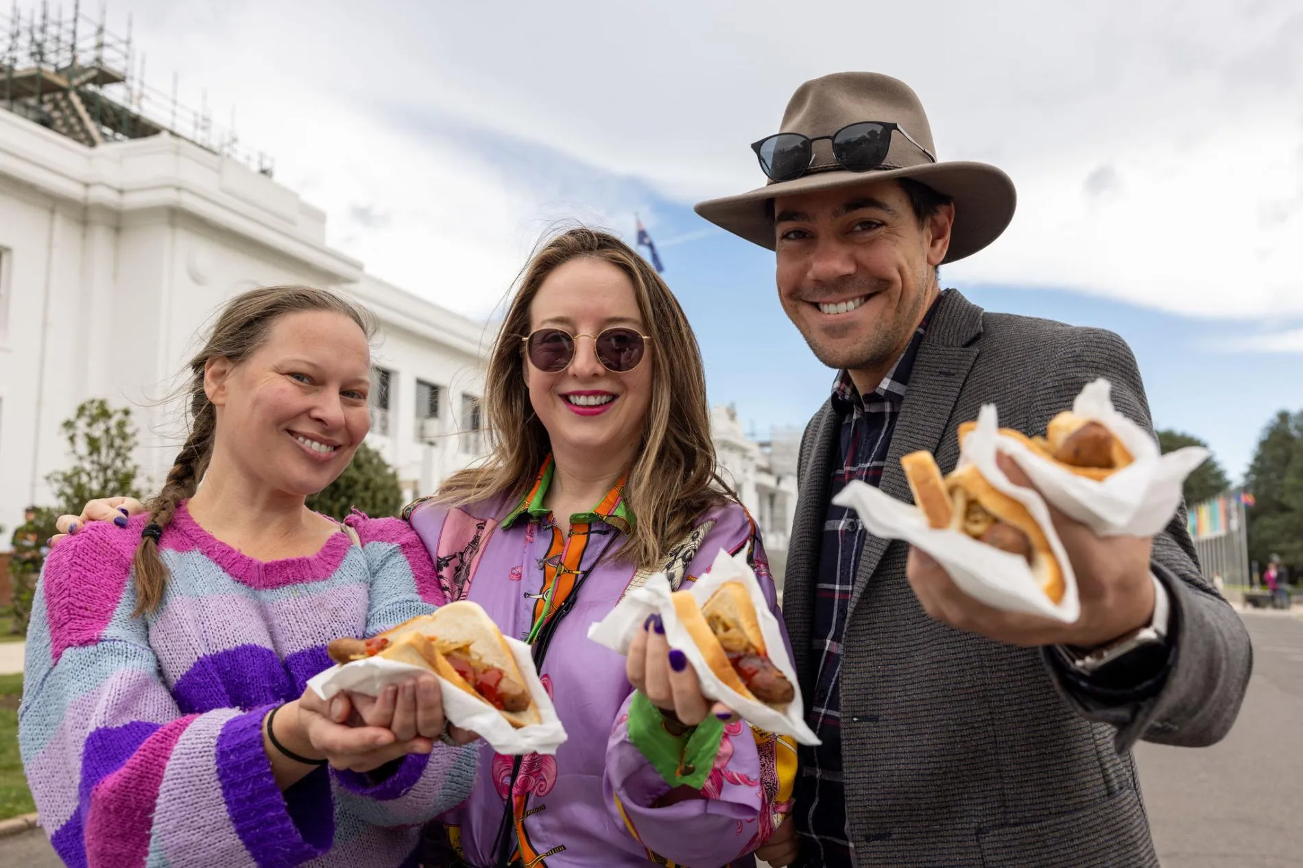 Two women wearing brightly coloured purple tops and a man in a jacket and hat stand in front of Old Parliament House holding sausage sandwiches.