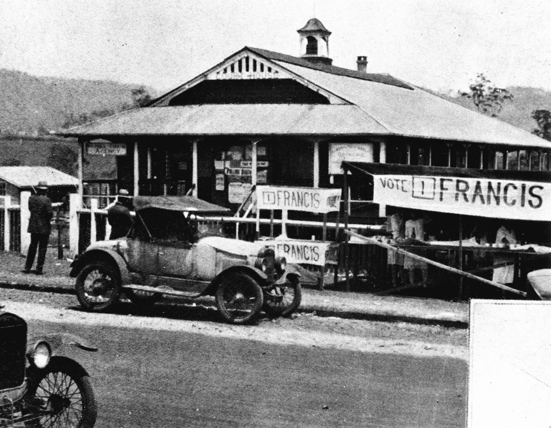 An old fashioned car on a street outside the old Atherton Courthouse in Queensland in 1928. Banners out the front read '1 Francis'. Women sell refreshments out the front at a stall.