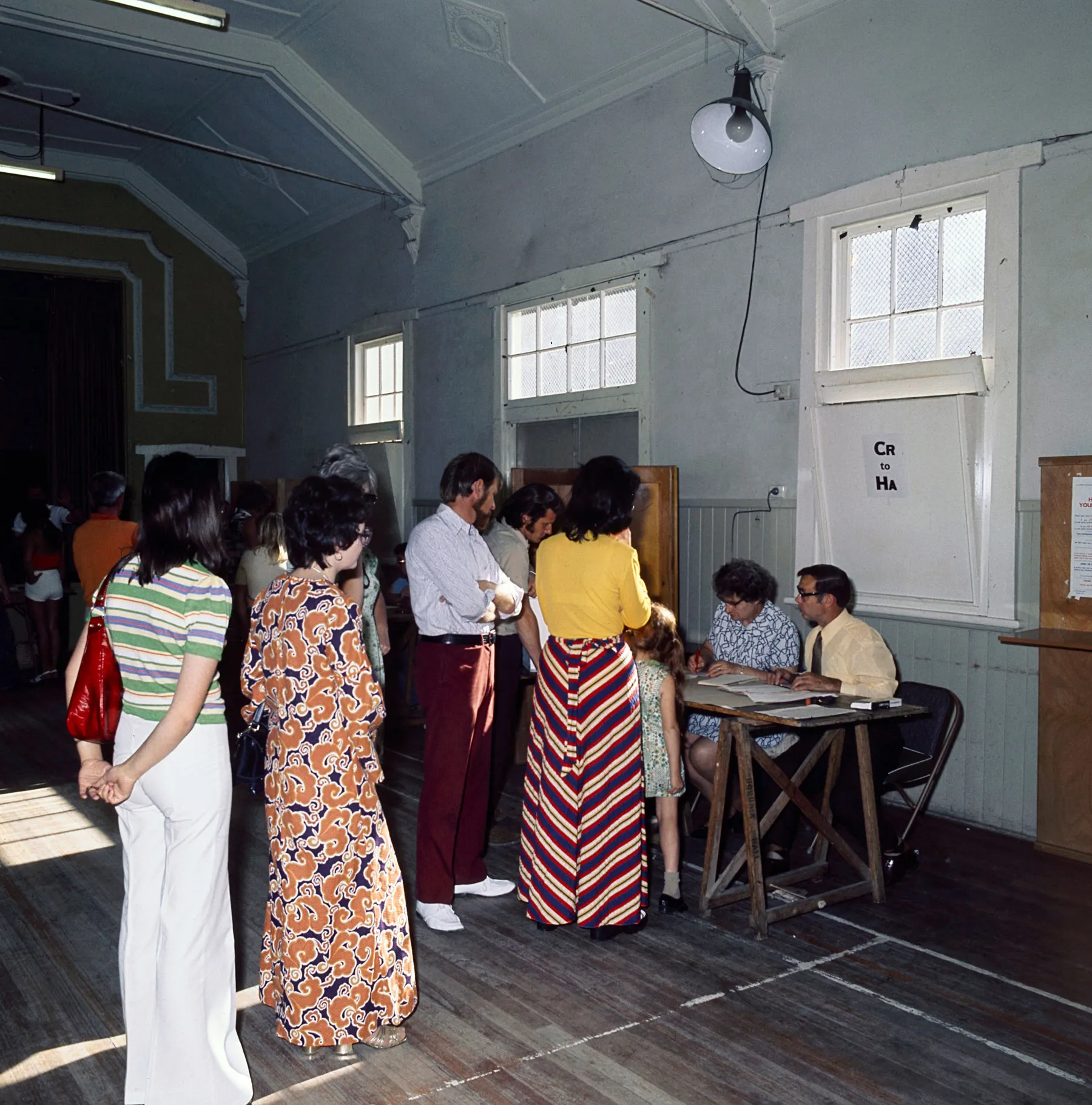 Men and women in colourful '70s clothes queue in a hall being used as a polling station. Election officials sit at wooden tables.