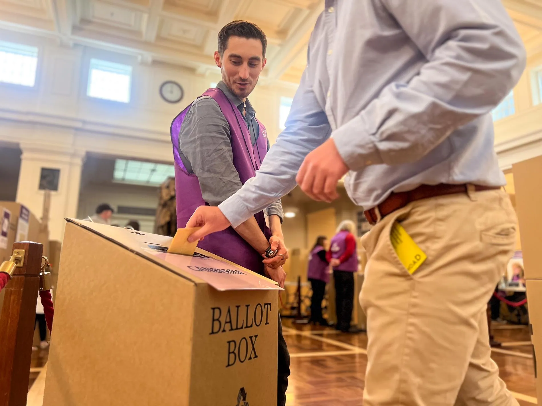 A voter puts a ballot paper in a cardboard ballot box while an AEC official dressed in a purple AEC vest looks on. They are in King's Hall in Old Parliament House.