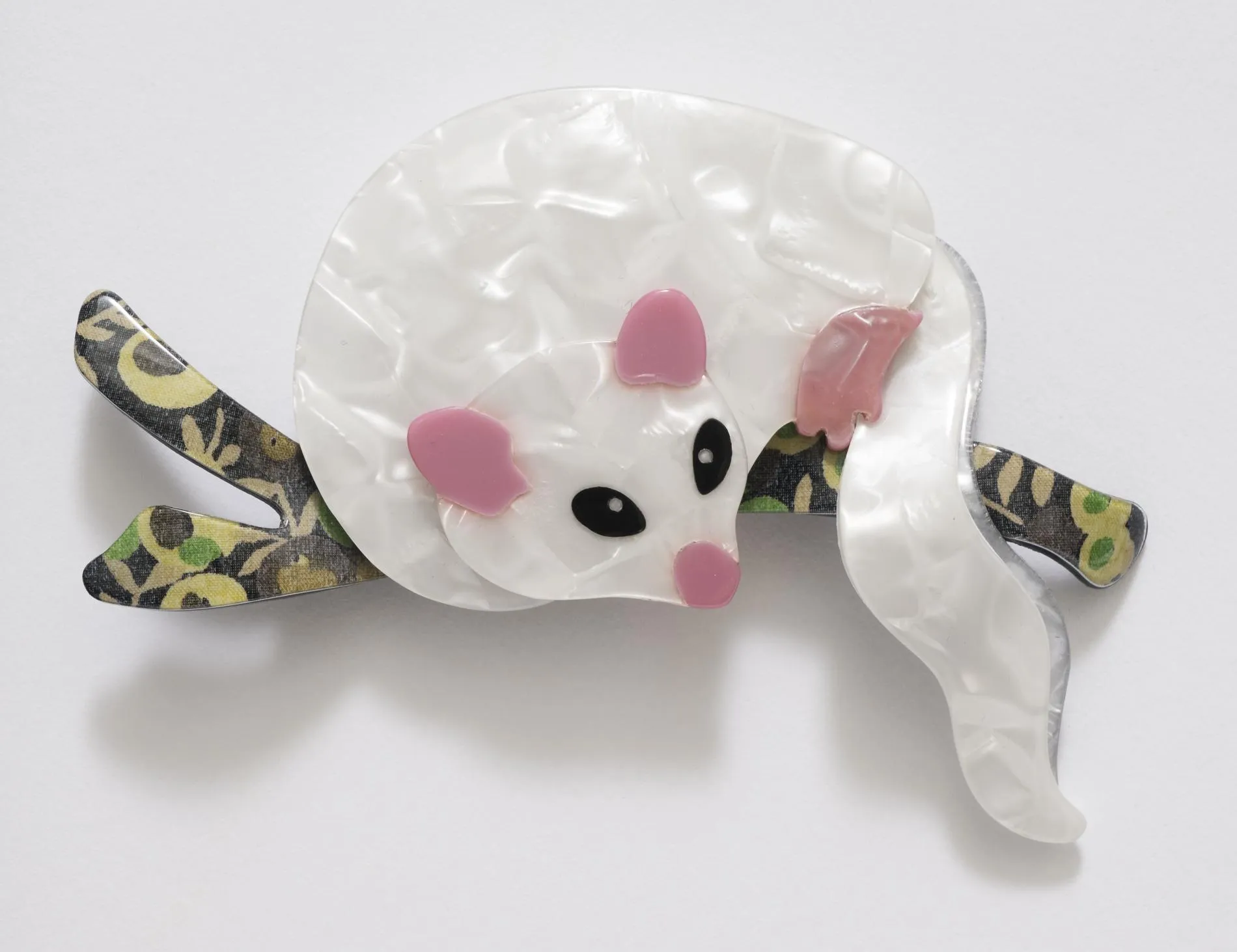 A celluloid possum brooch. The possum is white with pink ears, nose and feet, it sits on a black, green and gold branch.