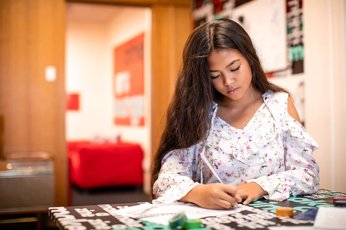A girl in a white floral top with brown hair writes with a pencil at a table in Old Parliament House.