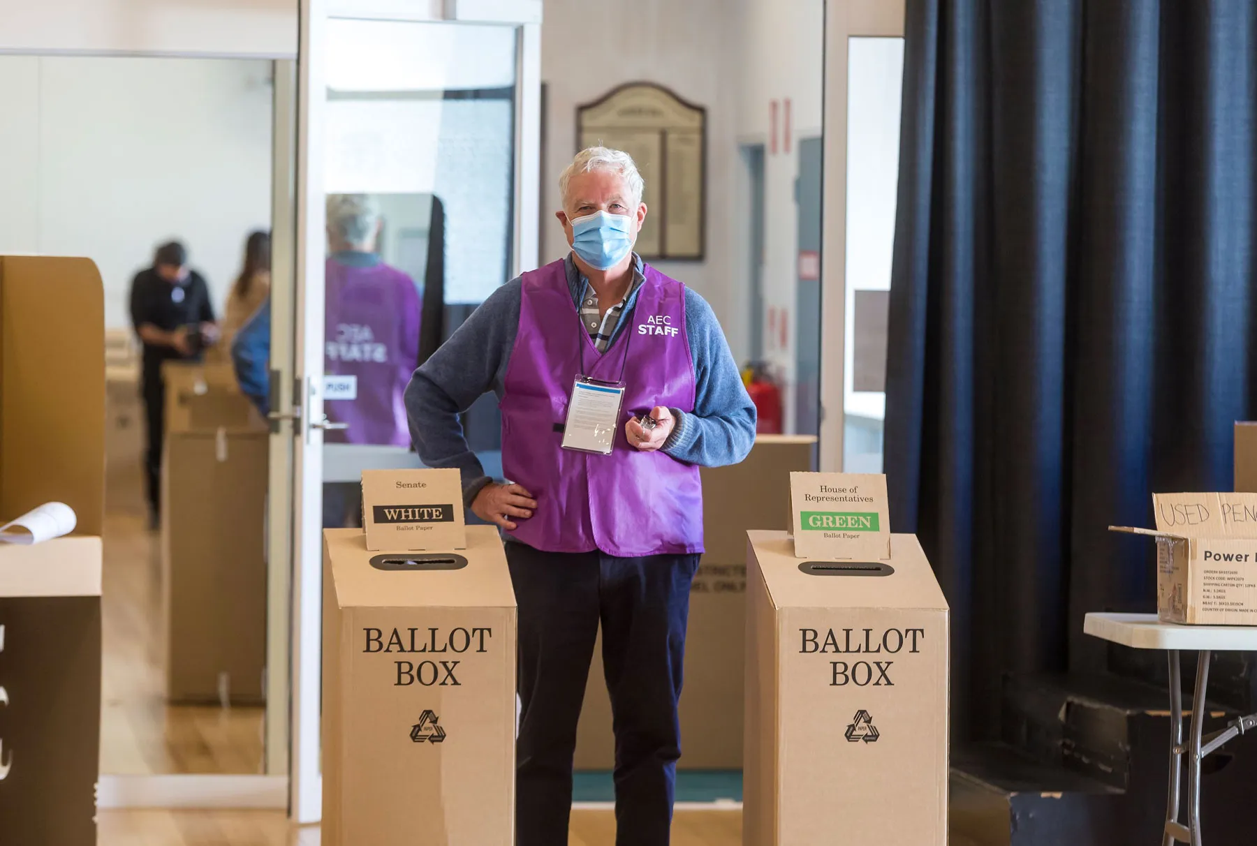 A man in a purple Australian Electoral Commission staff vest stands between two cardboard ballot boxes.