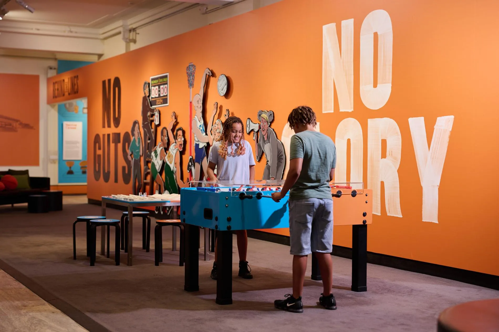 Two children play foosball in front of an orange wall with large graphic details that read No Gut, No Glory.