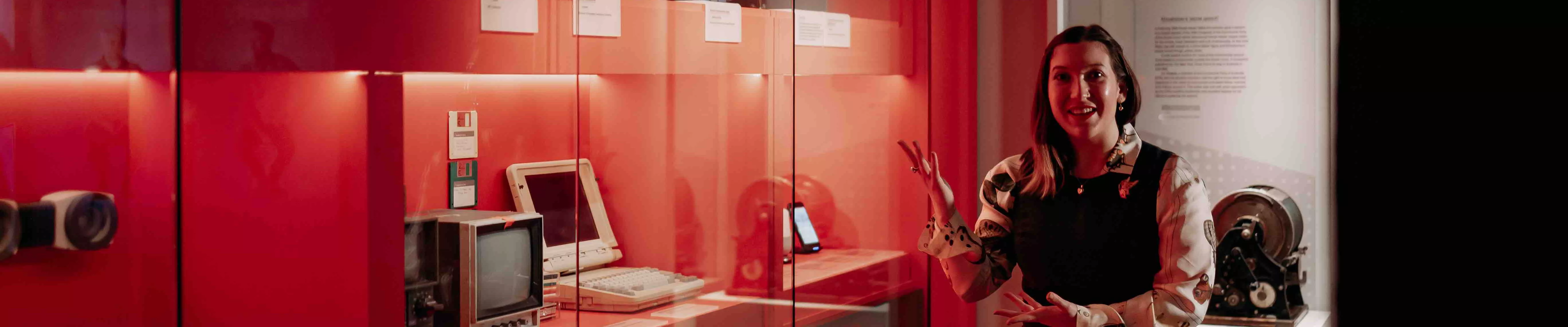 A MoAD educator stands in front of the Truth, Power and a Free Press exhibition wall featuring red walls with different types of media technology behind the glass case. 