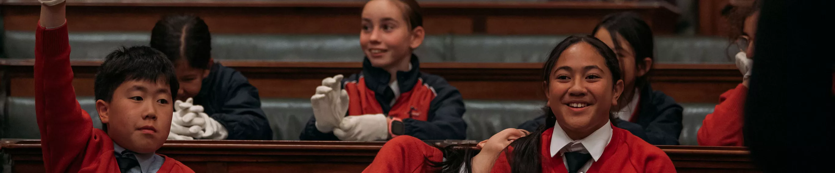 High school students in red jumpers sit on green leather seats in the House of Representatives Chamber. Two girls are laughing. 