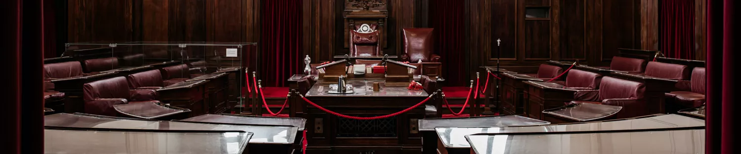 Two small steps with red carpet leading into the Senate Chamber with dark wooden furniture positioned in U-shape around a central table. 