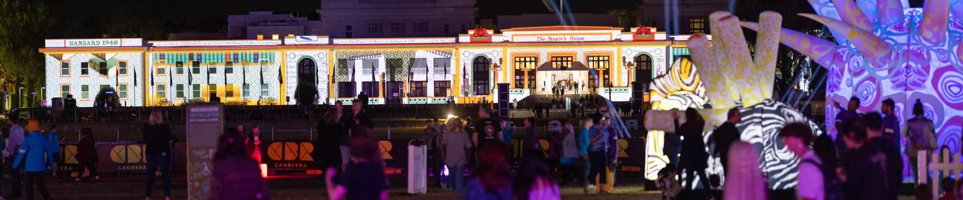 A night shot of Old Parliament House, a large white long building, with people gathering out the front and coloured lights projected onto the surface.