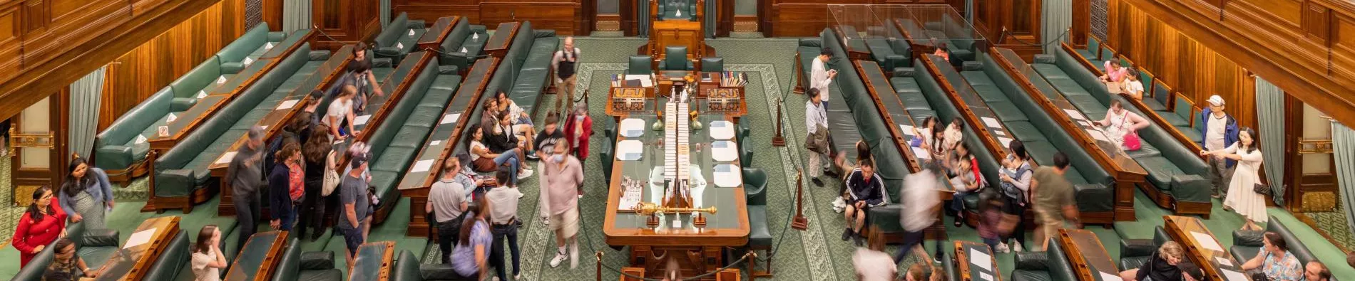 A view looking down onto the House of Representatives Chamber which has green carpet and green leather and timber benches in a U-shape surrounding a central table. 