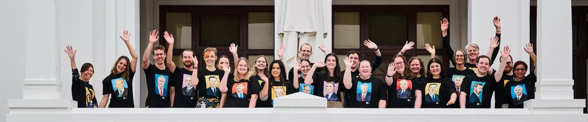 A group of people in black T-shirts with illustrations of prime ministers on them wave from a white terrace balcony.