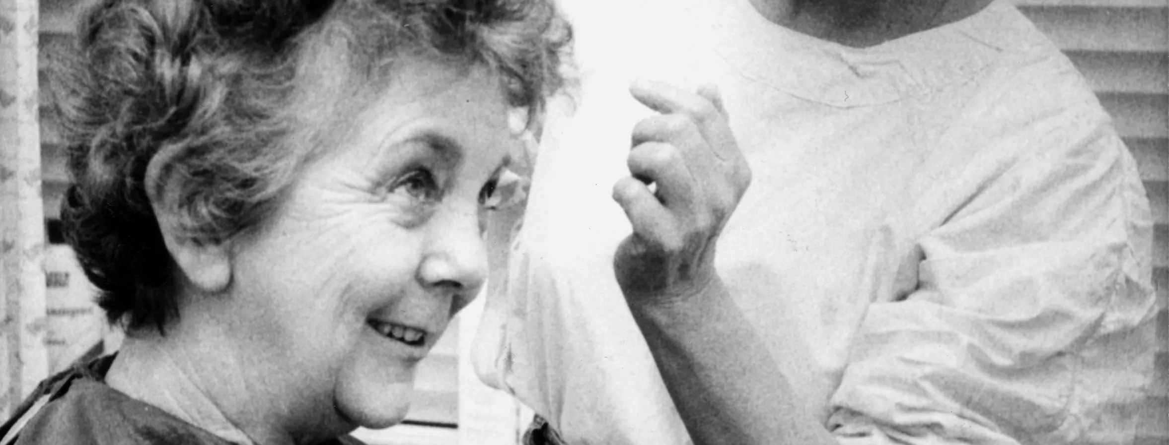 This black and white photograph depicts hairdresser Lizzie Scott, arranging Speaker Joan Child’s hair. Joan is sitting in the barber’s chair and is covered with a dark  cape while Lizzie stands to her left wearing a white shirt and jeans and holding a black comb.  