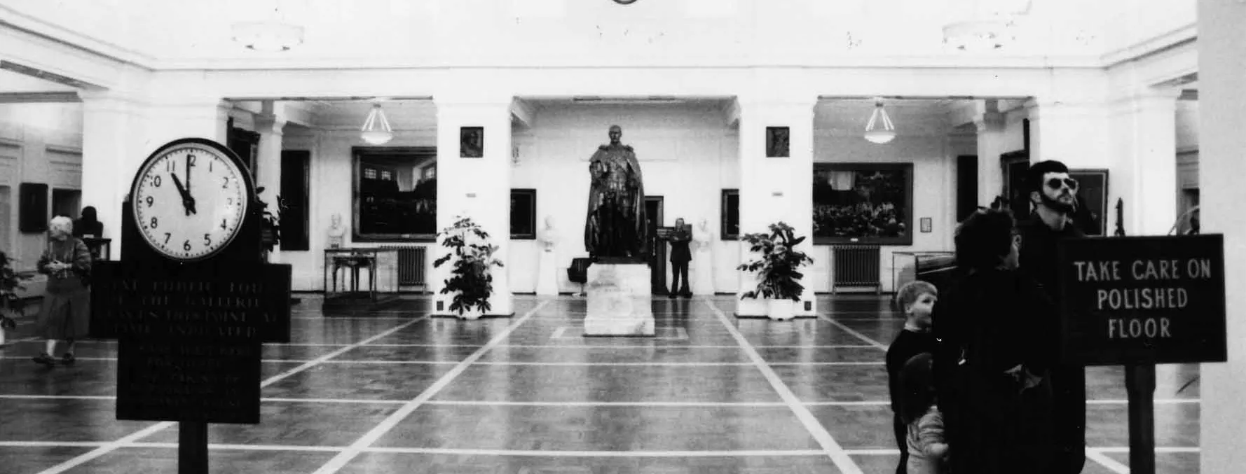 This black and white photograph of King’s Hall looks along the length of the room to the slightly larger than life bronze statue of King George the Fifth on a marble pedestal. King’s Hall is a large white room with a vaulted ceiling, celerestory windows, colonnades and a polished parquet floor.  The walls and columns feature artworks and portraits. You can see people wandering through the space including a family with young children and an older lady. In the foreground are two signs.