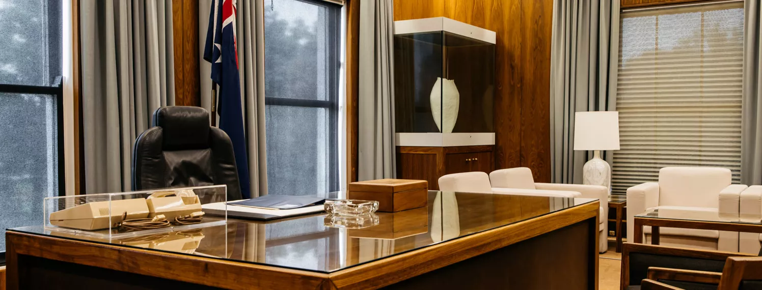 A colour photograph of the Prime Minister's Desk. Behind it is a comfortable office chair and an Australian flag on a pole. In front and to the side of the desk are armchairs.