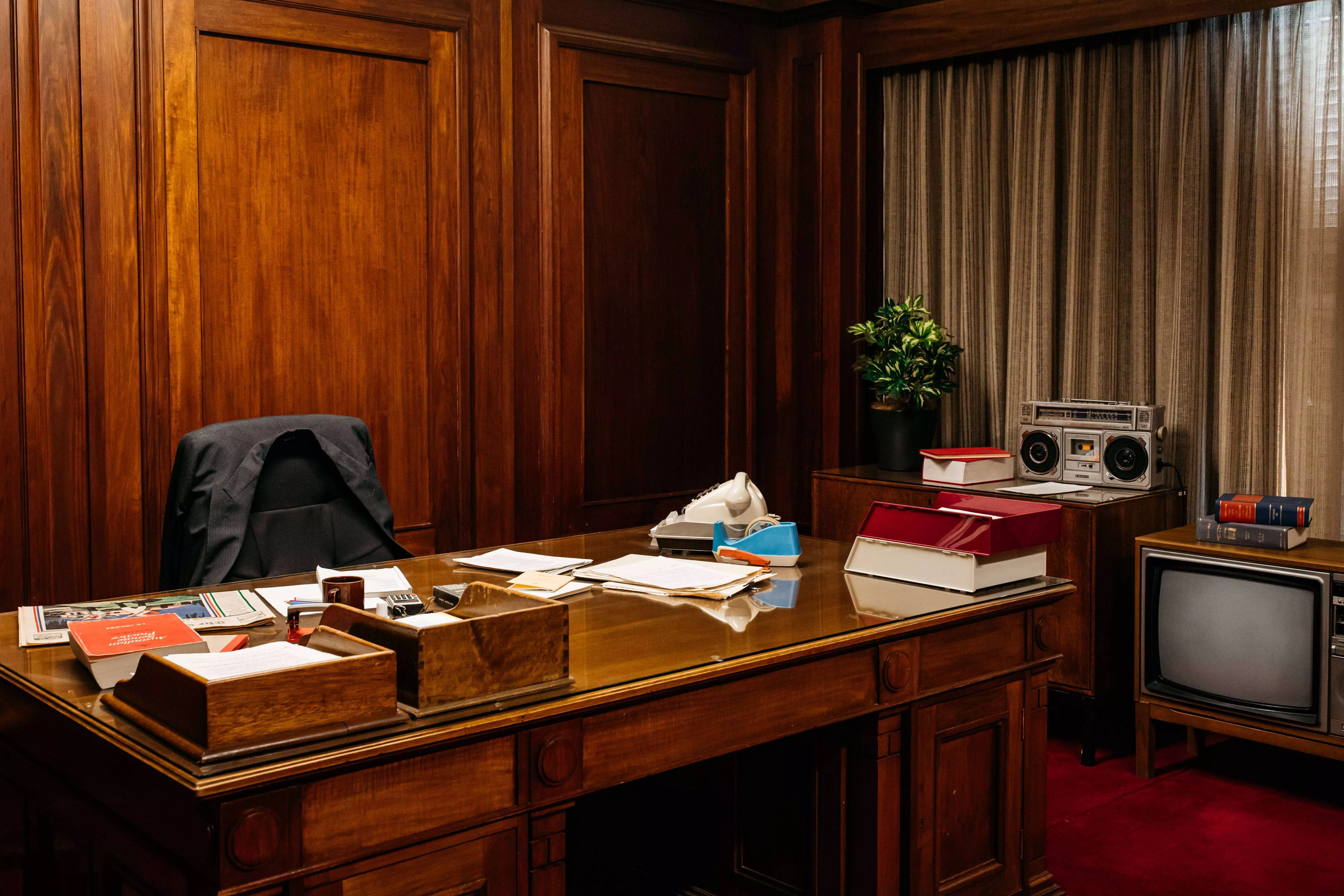 A dark timber panelled room with heavy curtins. A large wooden desk sits in the centre with papers, a telephone and timber trays. A stereo and old style TV sit in the corner. There is red carpet on the floor. A chair with a coat jacket sits at the desk. 