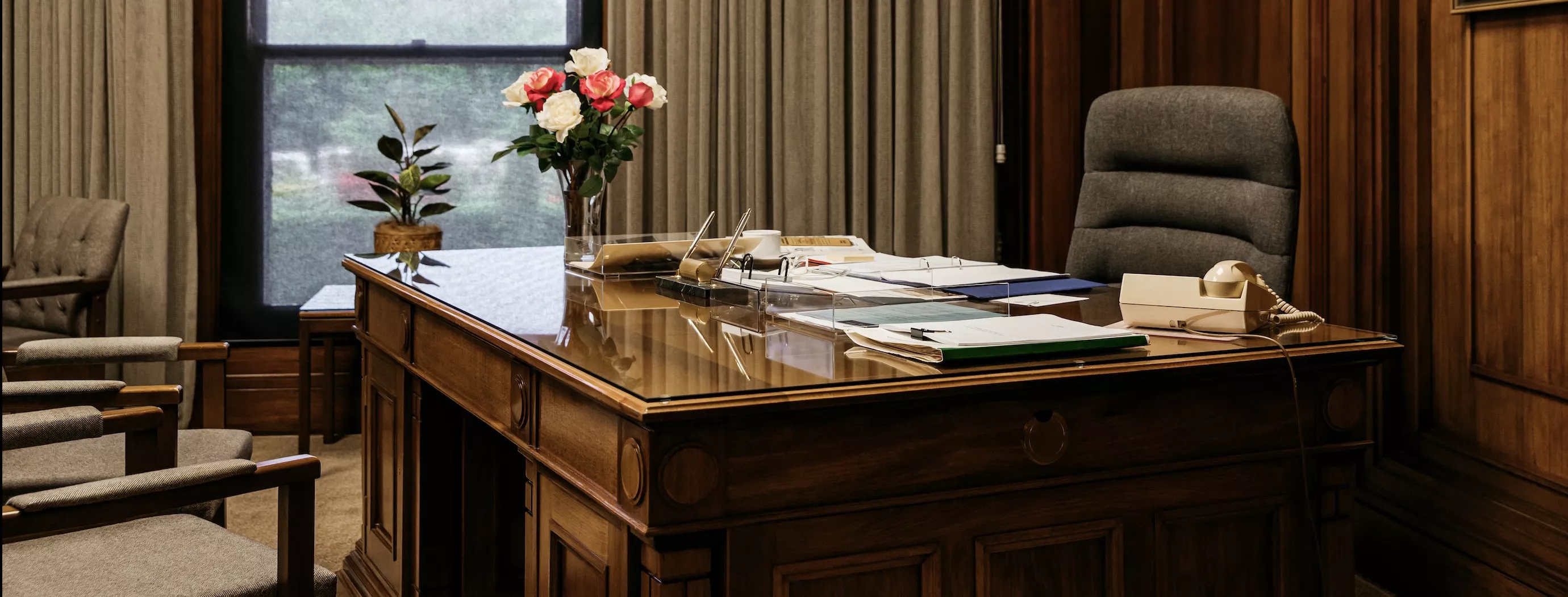 A large timber desk covered in folders and notes, with a vase with roses on it. 