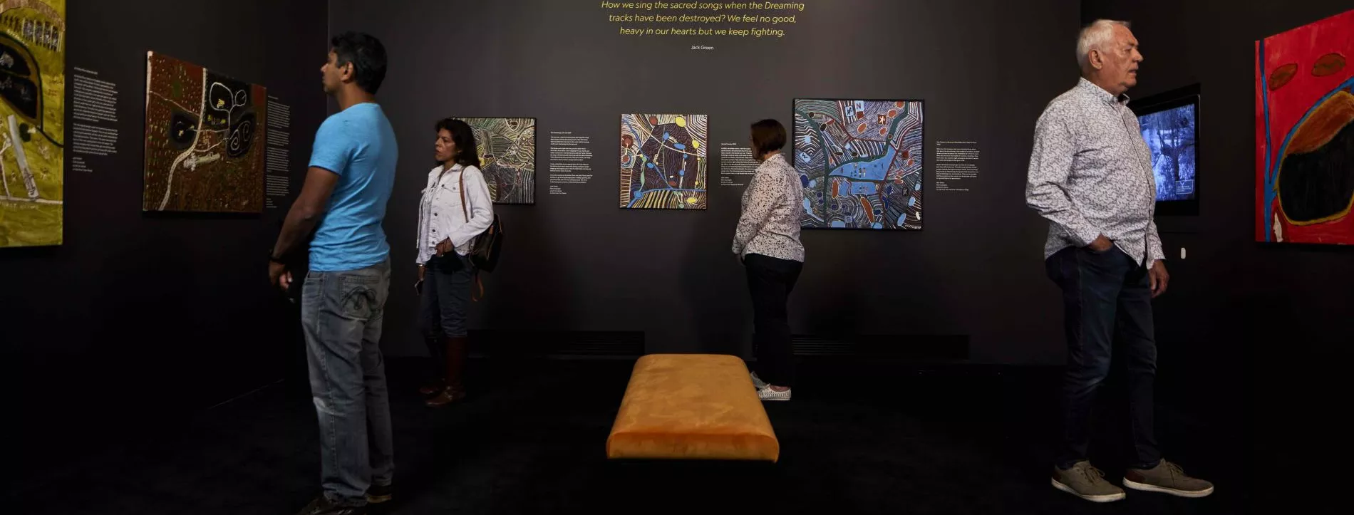 4 people stand in a museum room looking at paintings on the wall by artist Jack Green.