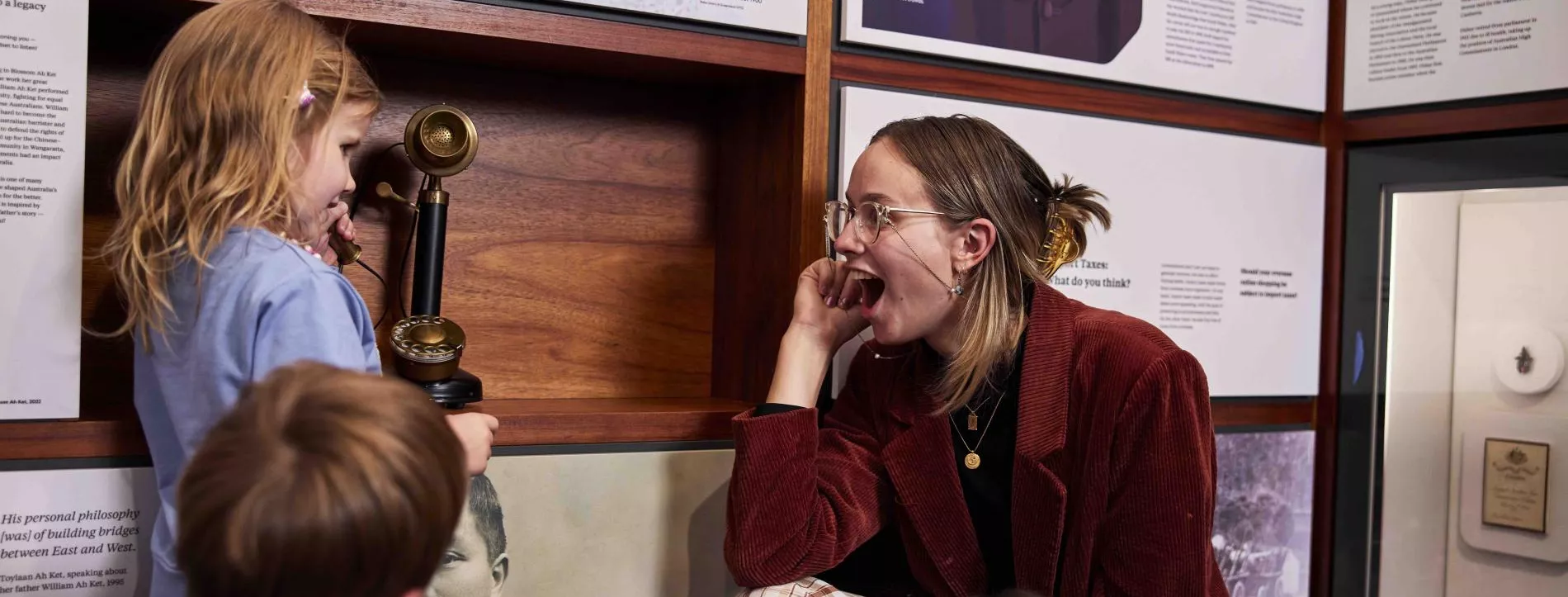 A woman smiles at a child holding a telephone to her ear in a museum exhibition with panels about prime ministers. 