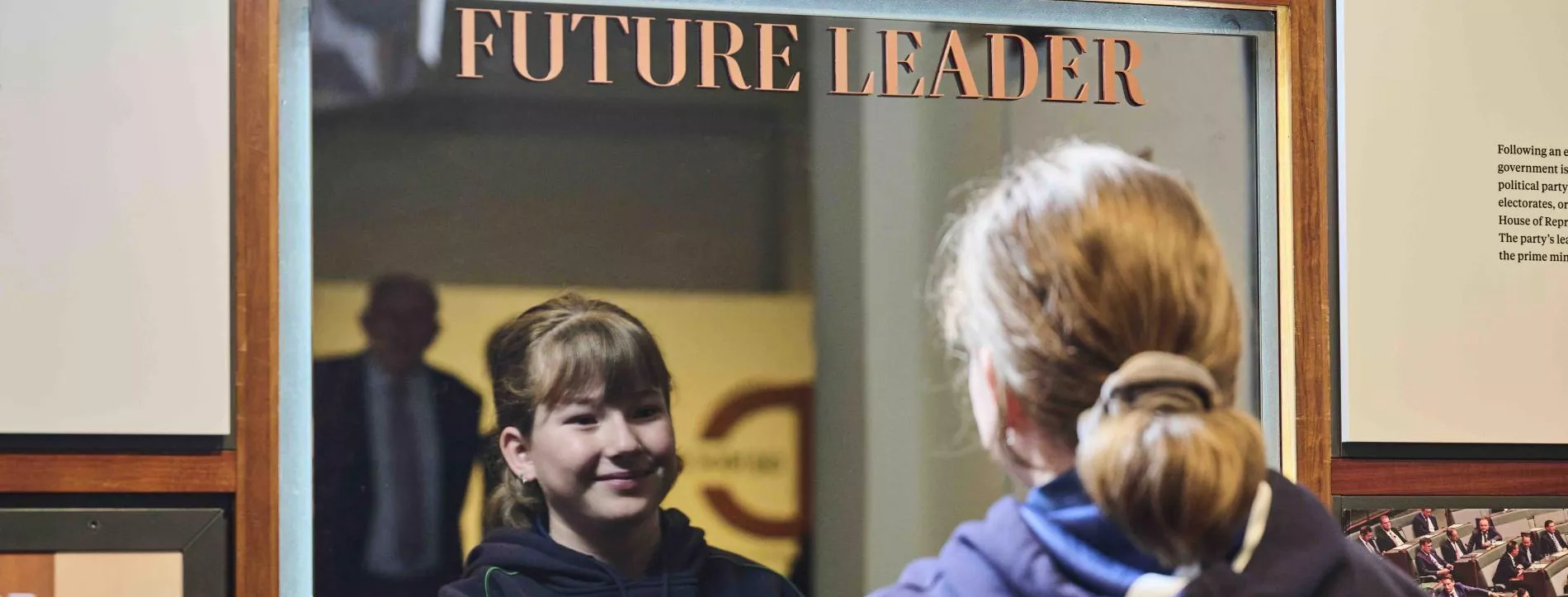 A primary school girl wearing her school jumper looks into a mirror with the words future leader on it. 