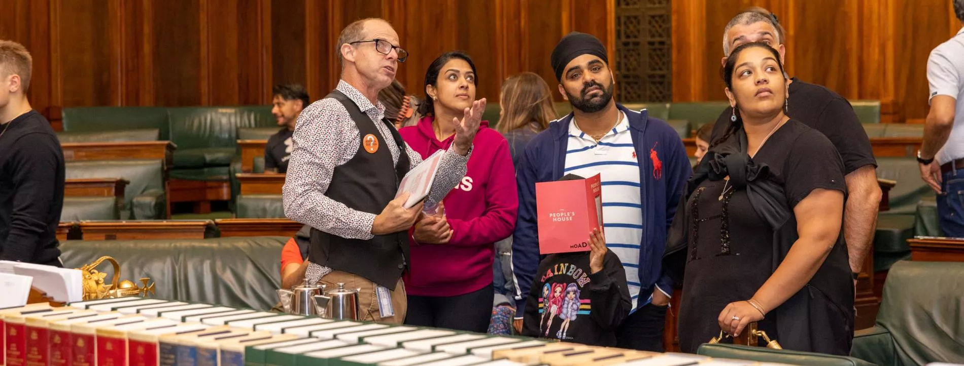 A tour guide points to something above a group of people listening to his tour. They are in the House of Representatives Chamber with green leather chairs behind them and leather bound books stacked on a central table in front of them.