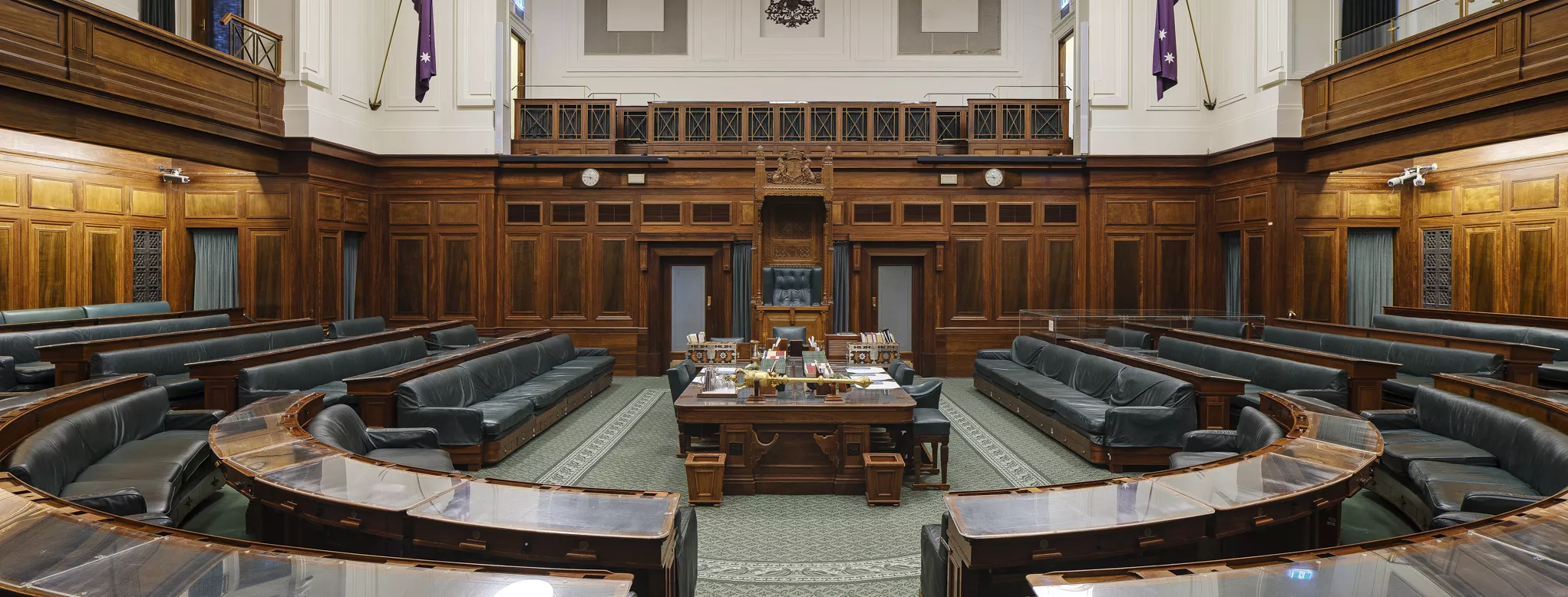 A green carpeted room with rows of wooden benches in a U shape facing a central table and the large, ornate Speaker's Chair. 