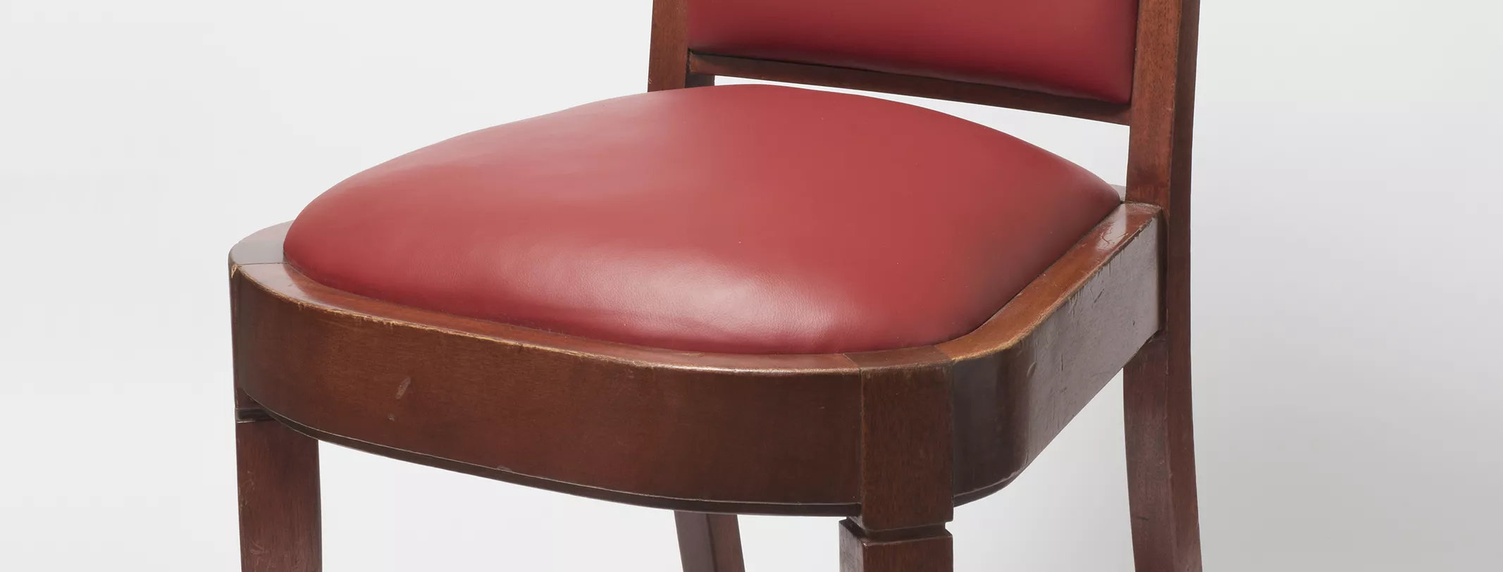 A wooden chair with cherry red leather seat cushion and back, and square legs with protective brass caps on the base of each leg. 