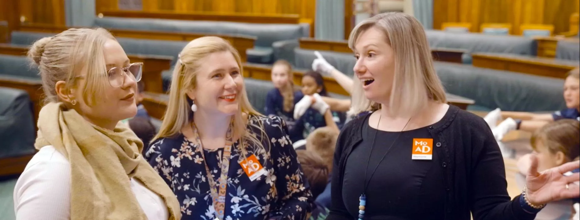 Two people stand holding clipboards, smiling and talking to a person about old parliament house's chamber of representatives.