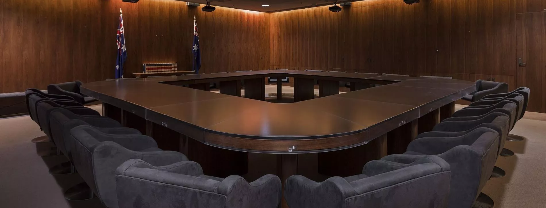 A dark room with heavy curtins and a large, heavy oval shaped table with a hole in the middle surrounded by large grey chairs. 