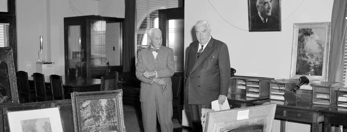 This black and white photograph shows two suited men standing side by side in the Government Party Room in 1962. On the left is Sydney businessman and philanthropist SH Ervin with a jaunty bow tie, on the right a more conservatively dressed Prime Minister Bob Menzies. They are surrounded by Australian paintings which are leaning against chairs, tables and walls. The two men are viewing the paintings that Ervin was donating to the National Collection. In the background are meeting tables and telephone booths