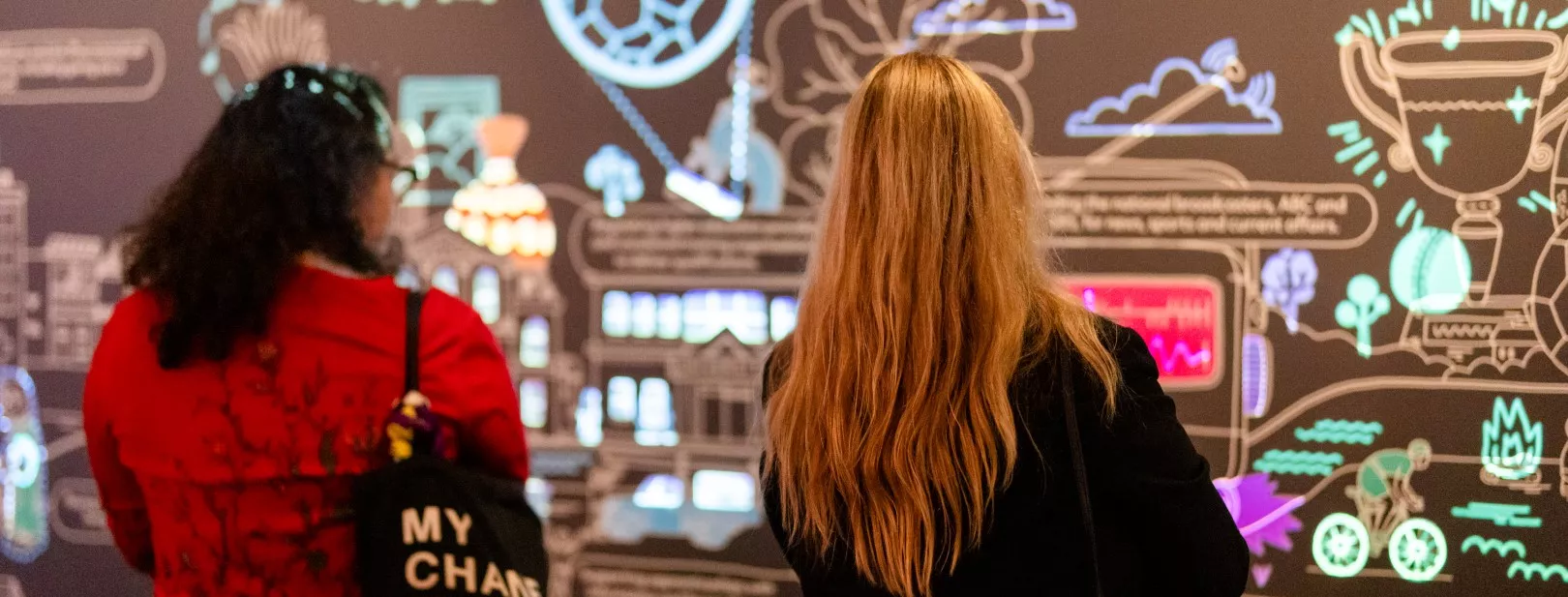 Two women look at a large wall installation of coloured lights and illustrations.