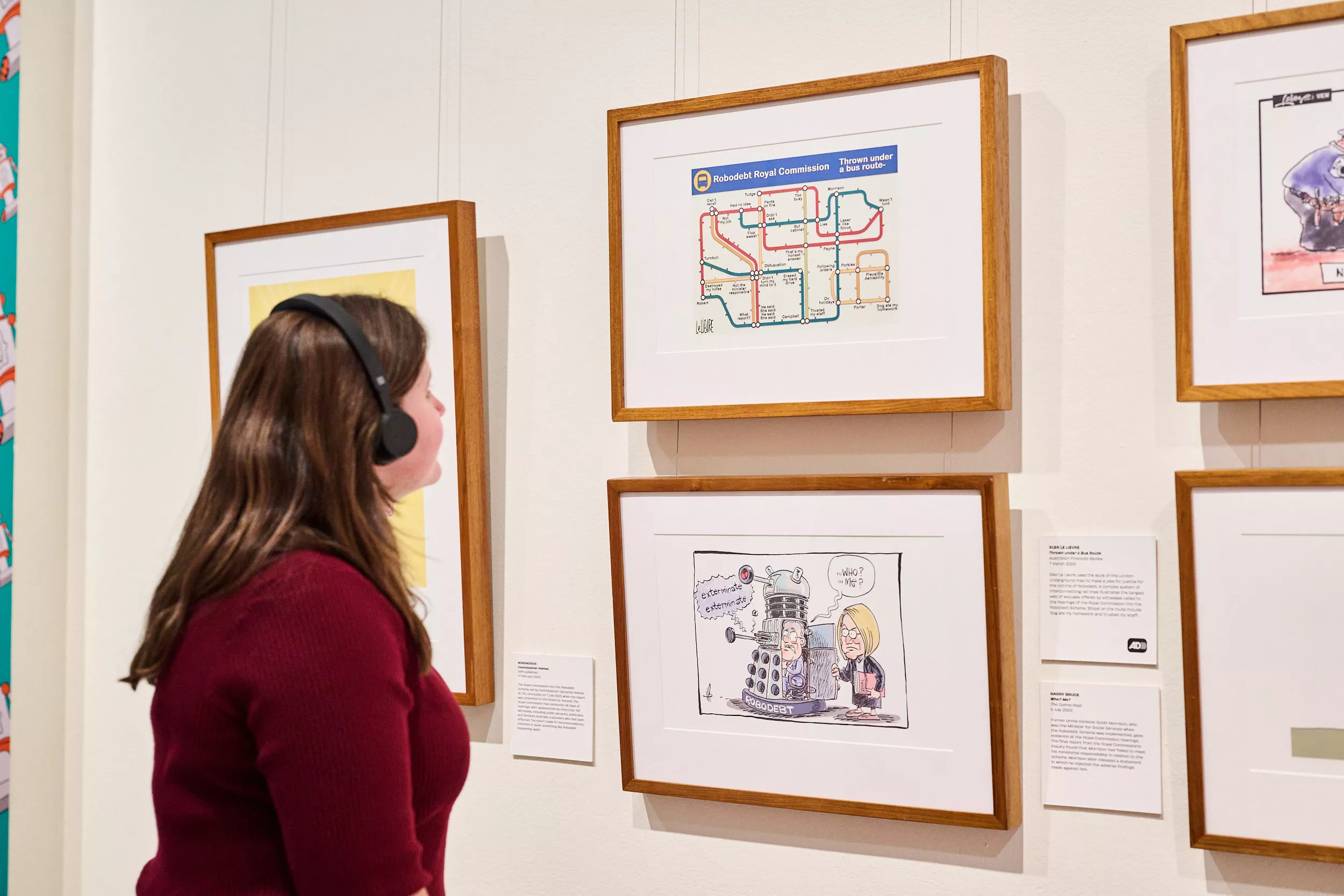 A woman wearing a dark red top and black headphones looks at framed cartoons with a caption featuring the Audio Description logo.