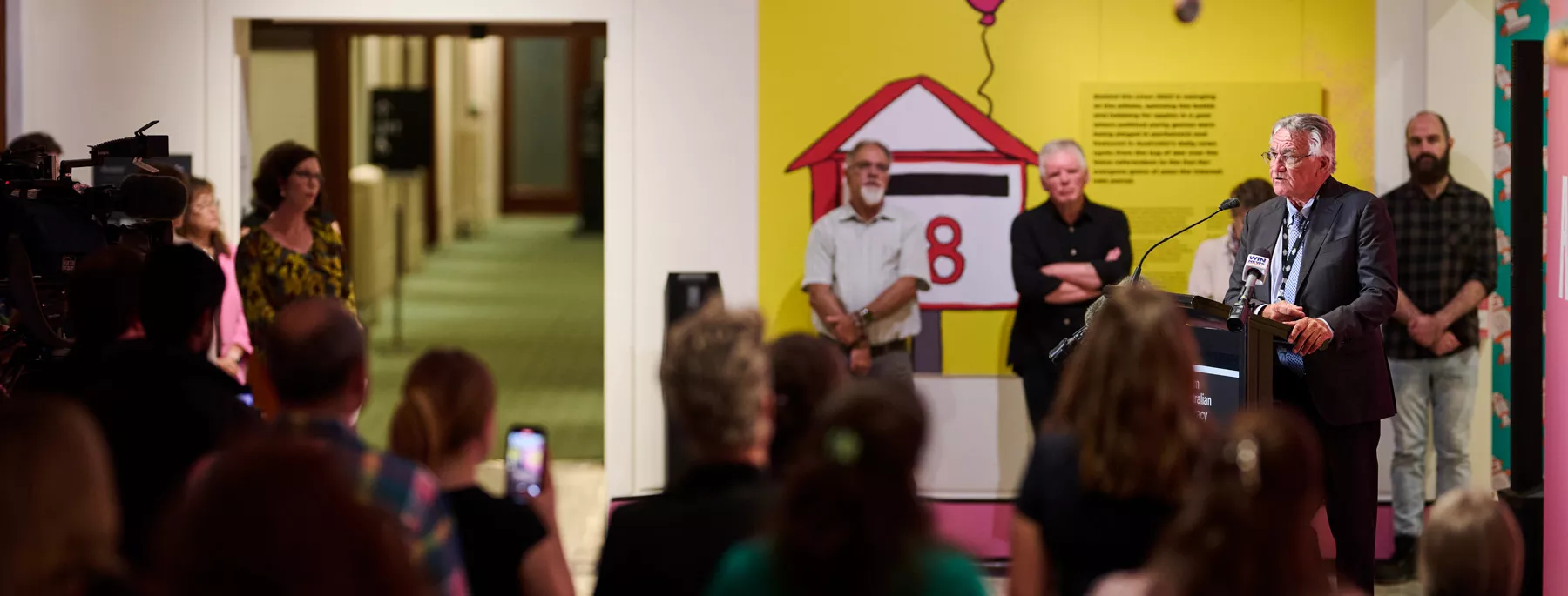 Barrie Cassidy stands behind a podium and speaks to an audience, some of whom are recording on photos and cameras. 