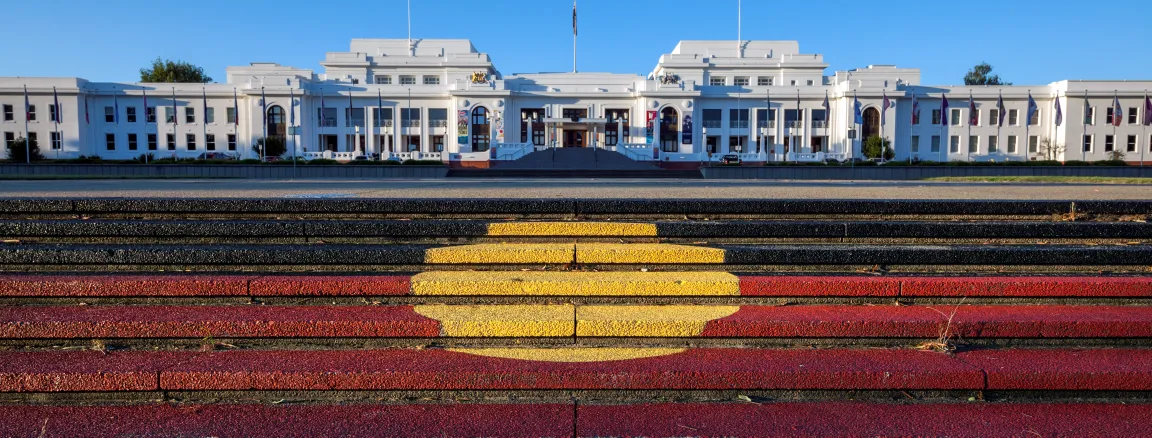 Steps with the Aboriginal flag painted on them lead up to a view of a large long white building, Old Parliament House.