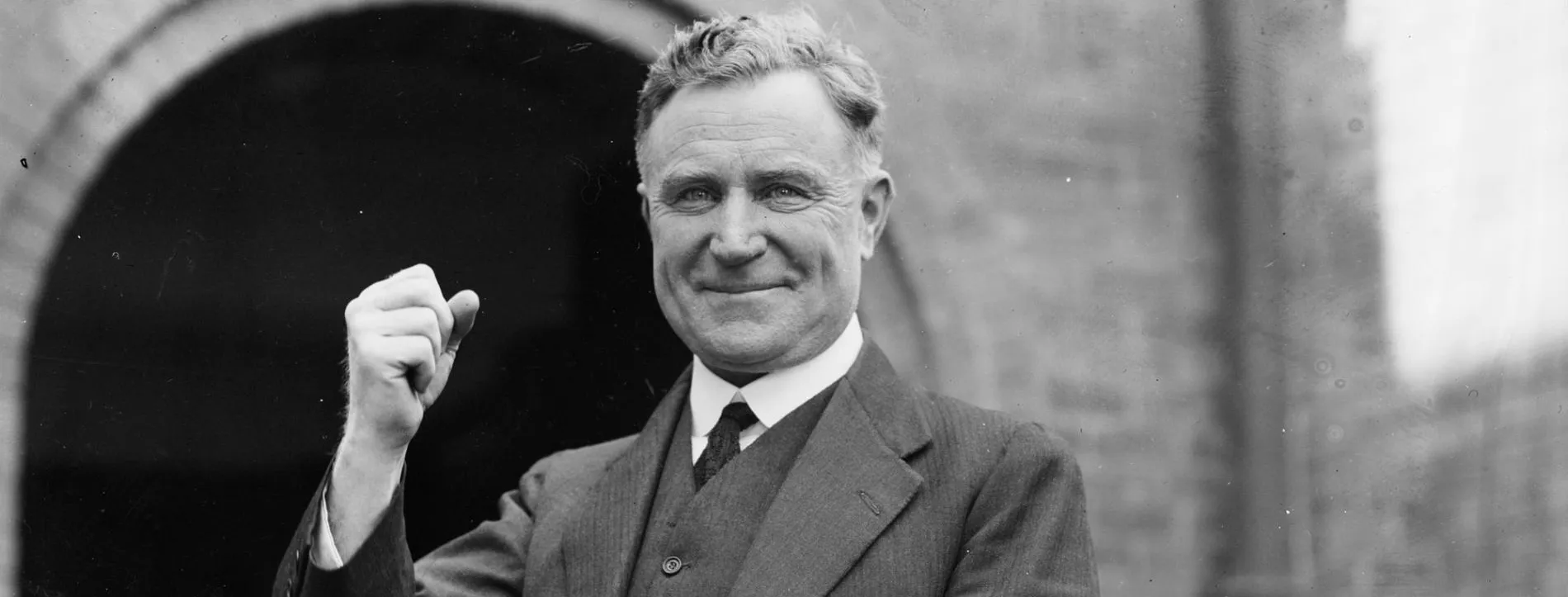 A man, Earle Page, wearing a striped three-piece suit, smiles and poses for a camera with a raised fist.