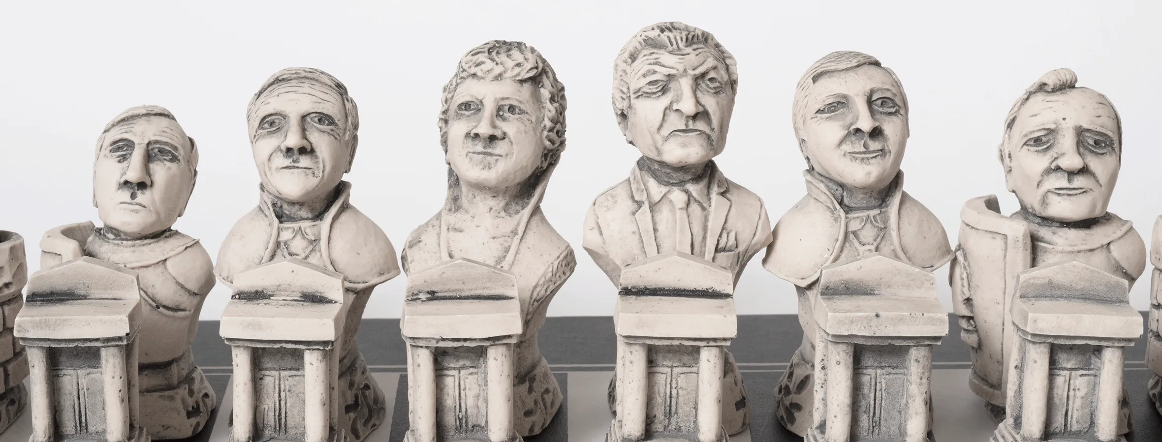 A close up of six chess pieces standing in a row on a black and white checkered chess board. Each chess piece is carved from white stone with sombre faces in the likeness of politicians.