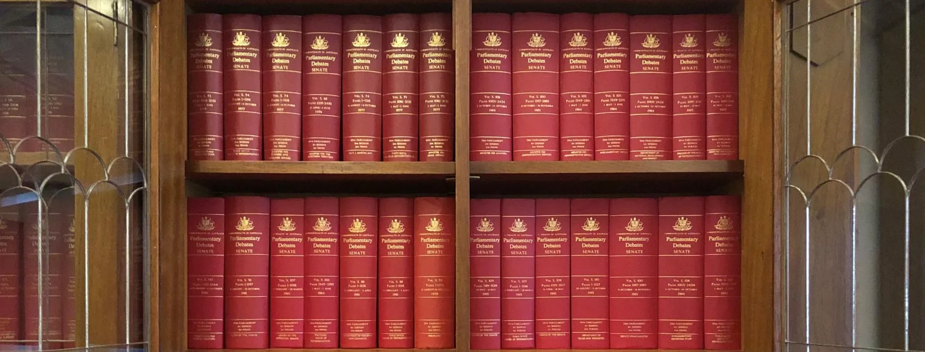 A wooden shelf with opened glass doors holds three rows of red bound volumes labelled 'Parliamentary Debates; Senate' on the spines.