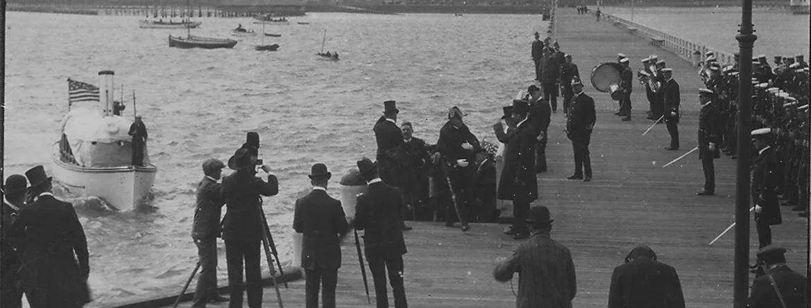 An elevated view from above and behind small clusters of people who stand on a wooden pier. They are looking out at a boat that is headed towards the pier.  The label reads "The American Fleet in Australia. Rear-Admiral Sperry meeting the Prime Minister (Hon. Alfred Deakin) at St. Kilda." 1908.