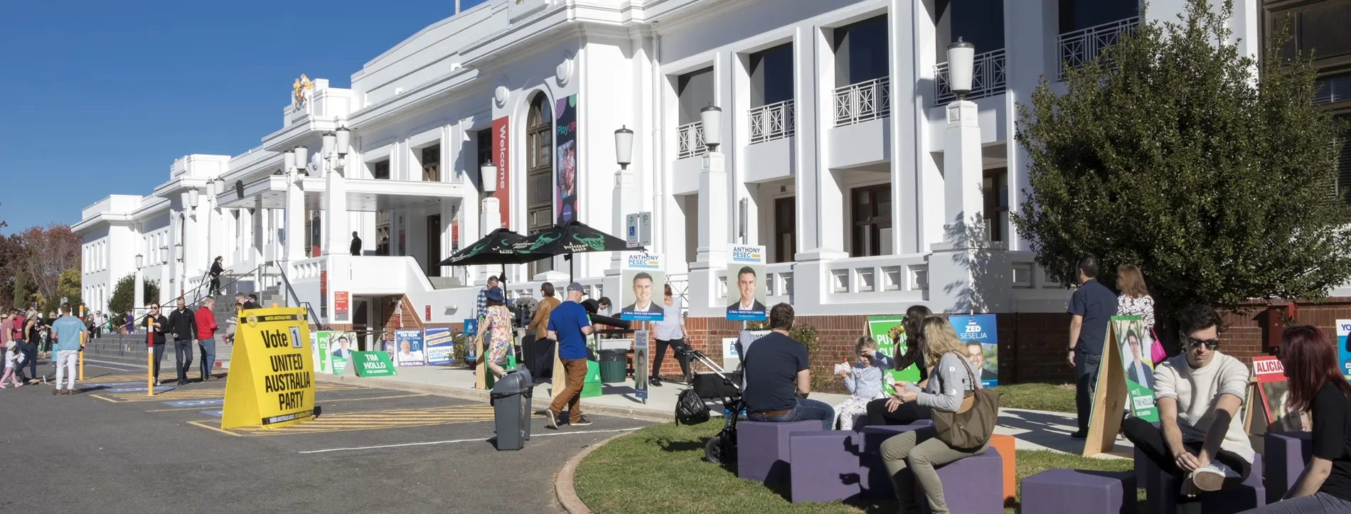 Many colourful signs and placards displaying political candidates, as people enter and leave Old Parliament House, with some sitting on purple blocks on the front lawn.