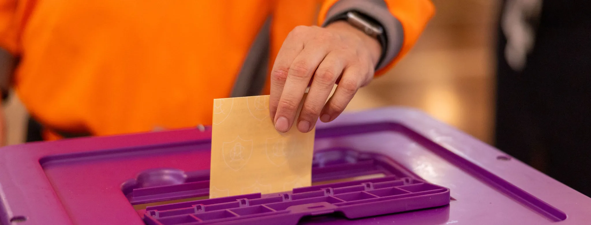 A person wearing an orange long-sleeved top posts a yellow voting ballot into a purple box.