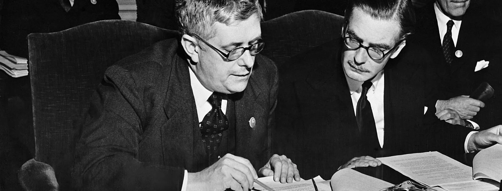 Dr Herbert Evatt sits next to Anthony Eden, reading through typed paper documents, at a table with a glass jug of iced water, and a nameplate labelled 'Australia'.